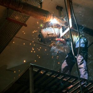 A welder working on metal pipes indoors, emitting bright sparks from the welding process.